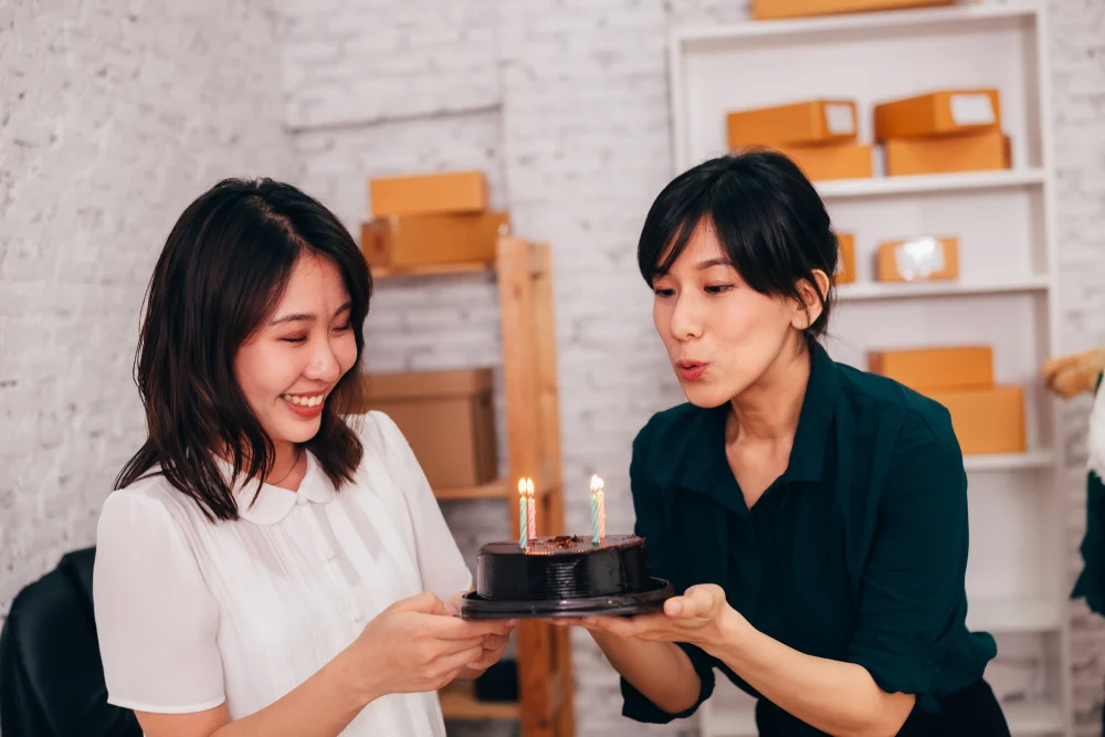 2 colleagues holding a chocolate cake and one of them is blowing the candle