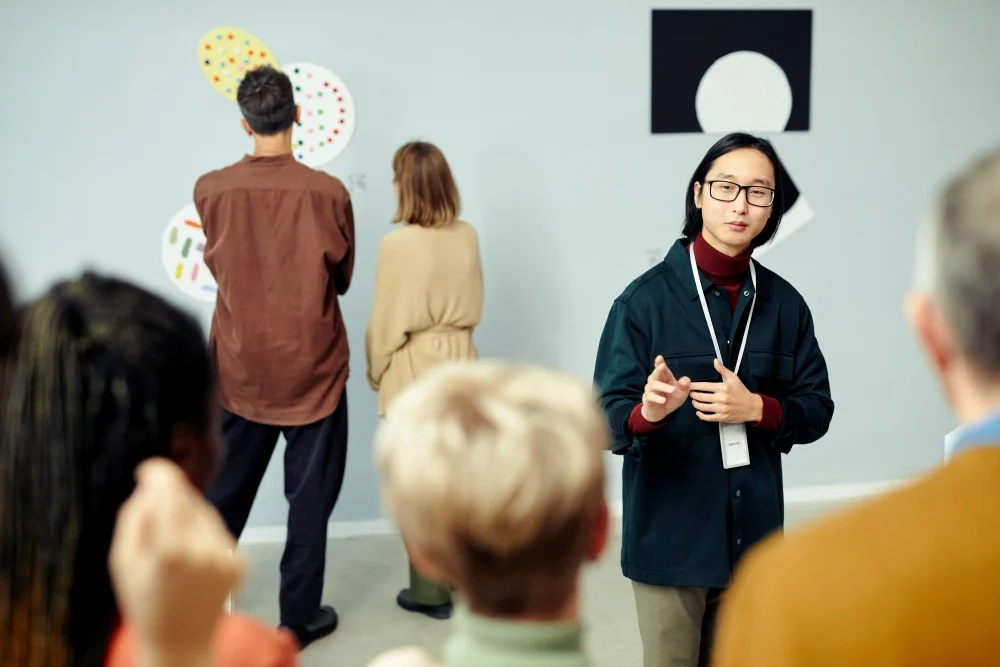 Young Asian gallerist standing in front of group of tourists speaking about contemporary artworks demonstrated at exhibition