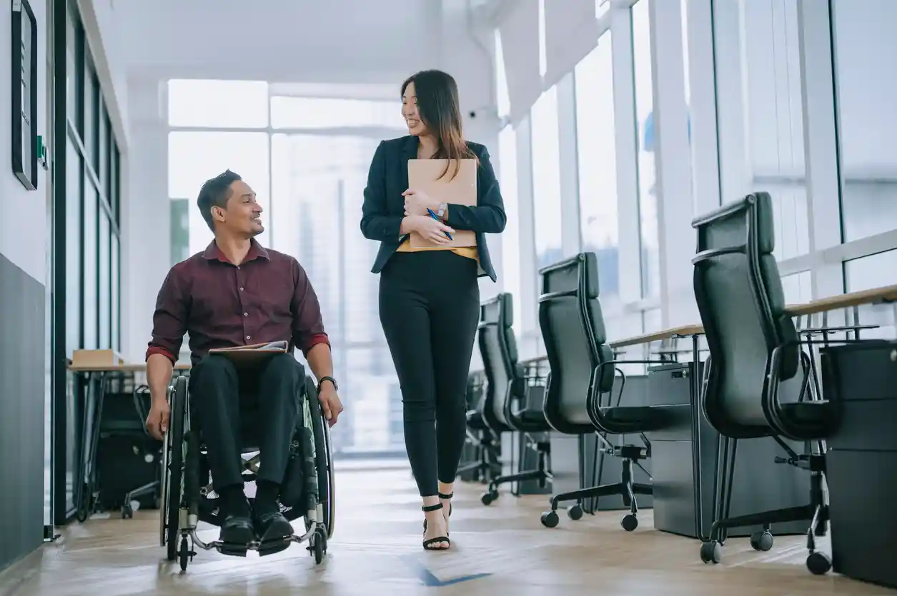Indian white collar male worker in wheelchair having cheerful discussion conversation with his female Asian chinese colleague coworking at walkway corridor