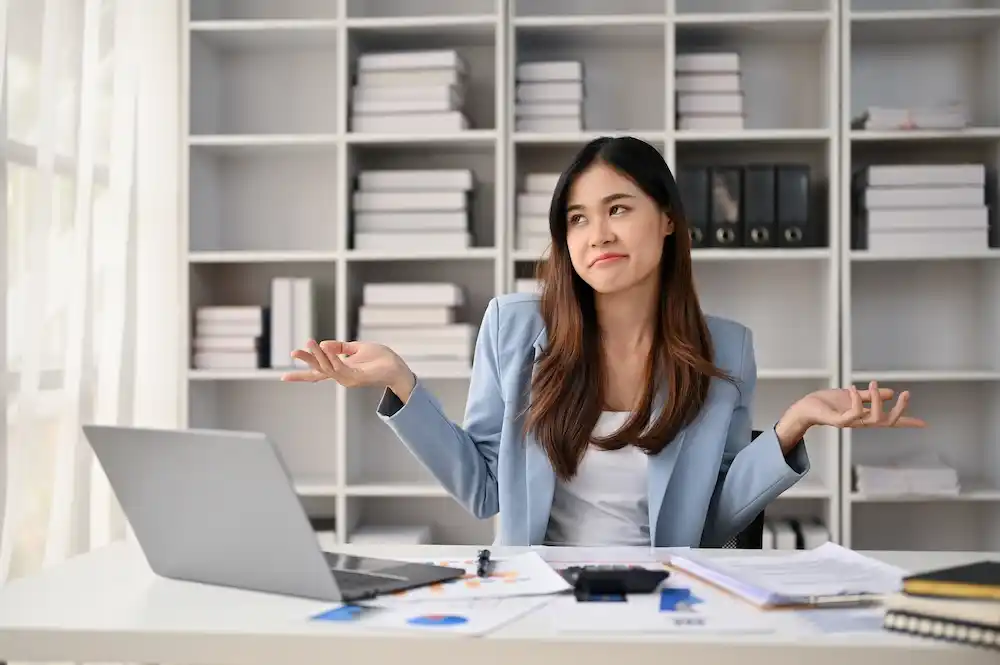 Millennial Asian professional sits at her office desk shrugging her shoulders
